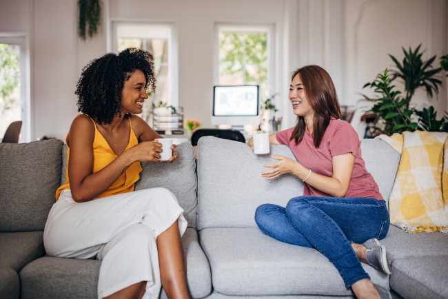 Two women sitting on sofa at home, they are drinking coffee and talking.