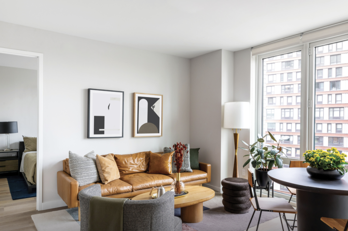 living room with brown leather sofa, white walls, & large windows looking out on another building