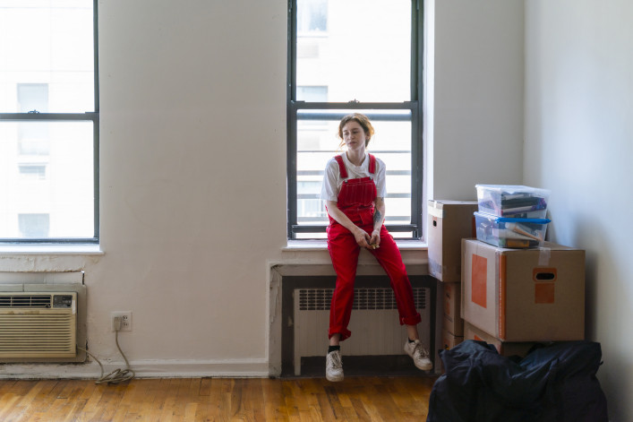 Young woman is sitting on a windowsill in an empty room of a newly rented apartment, tired after moving in. stock photo