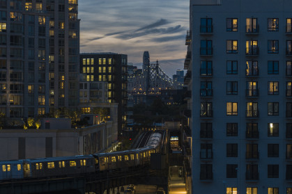 An elevated subway train riding on the line in Long Island City, Queens with Queensboro Bridge behind.