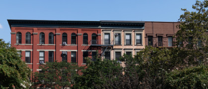 A row of colorful old brick apartment buildings surrounded by green trees during the summer in Harlem of New York City